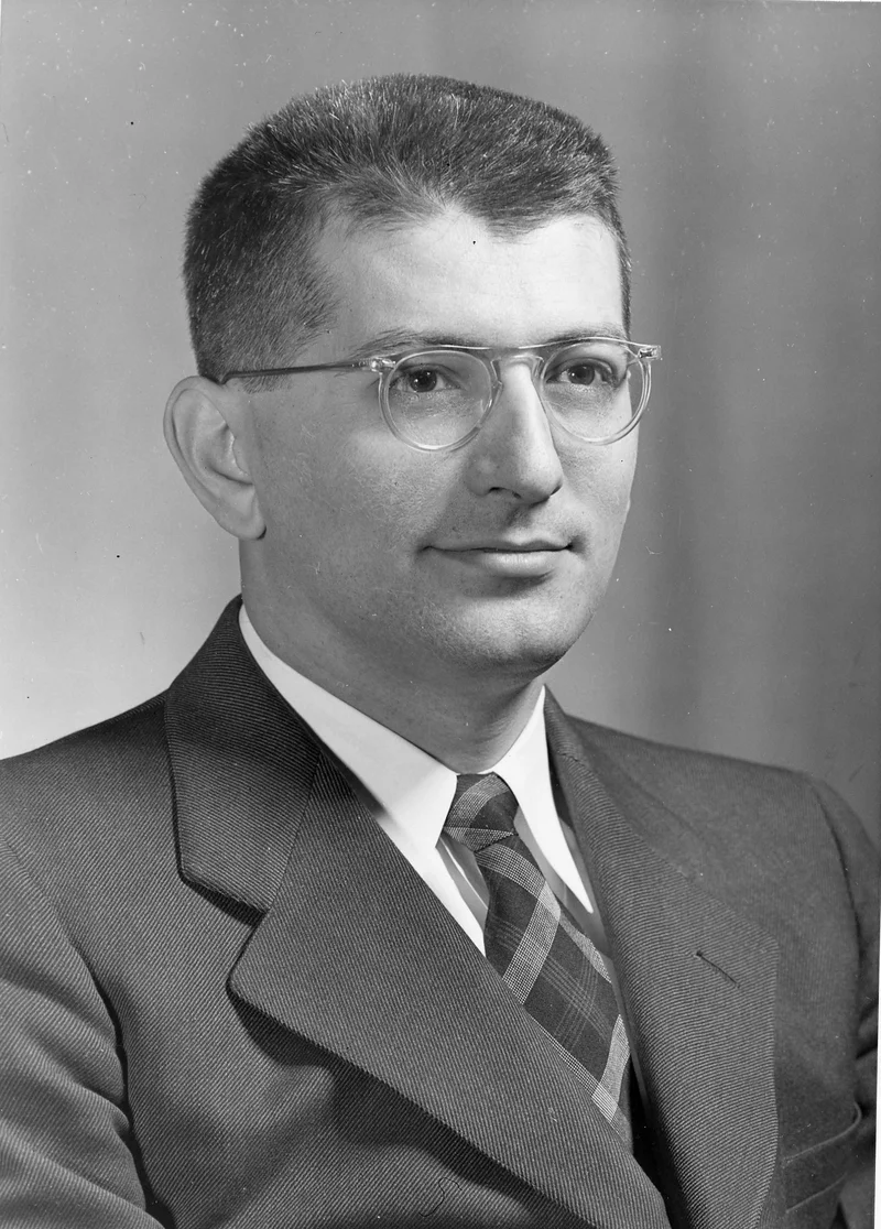 Studio portrait of Alex Bavelas in clear-framed glasses, a suit and a plaid tie